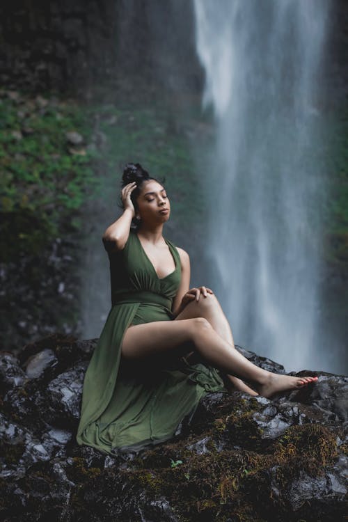 Photo of Woman in Green Dress Sitting on Rock Formation With Waterfall in the Background