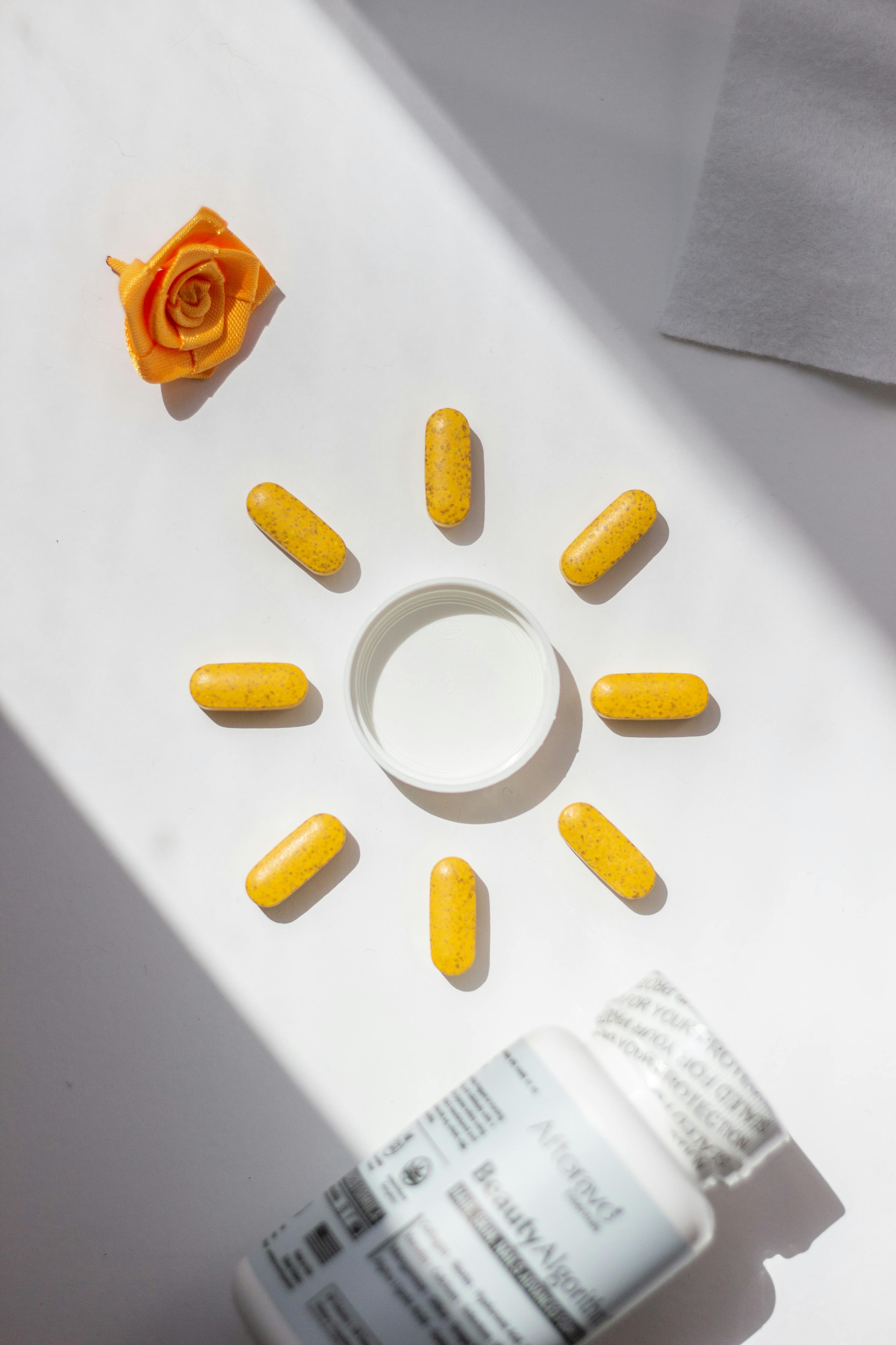 Bright yellow vitamin pills arranged in a sun shape with bottle and rose accent on white background.