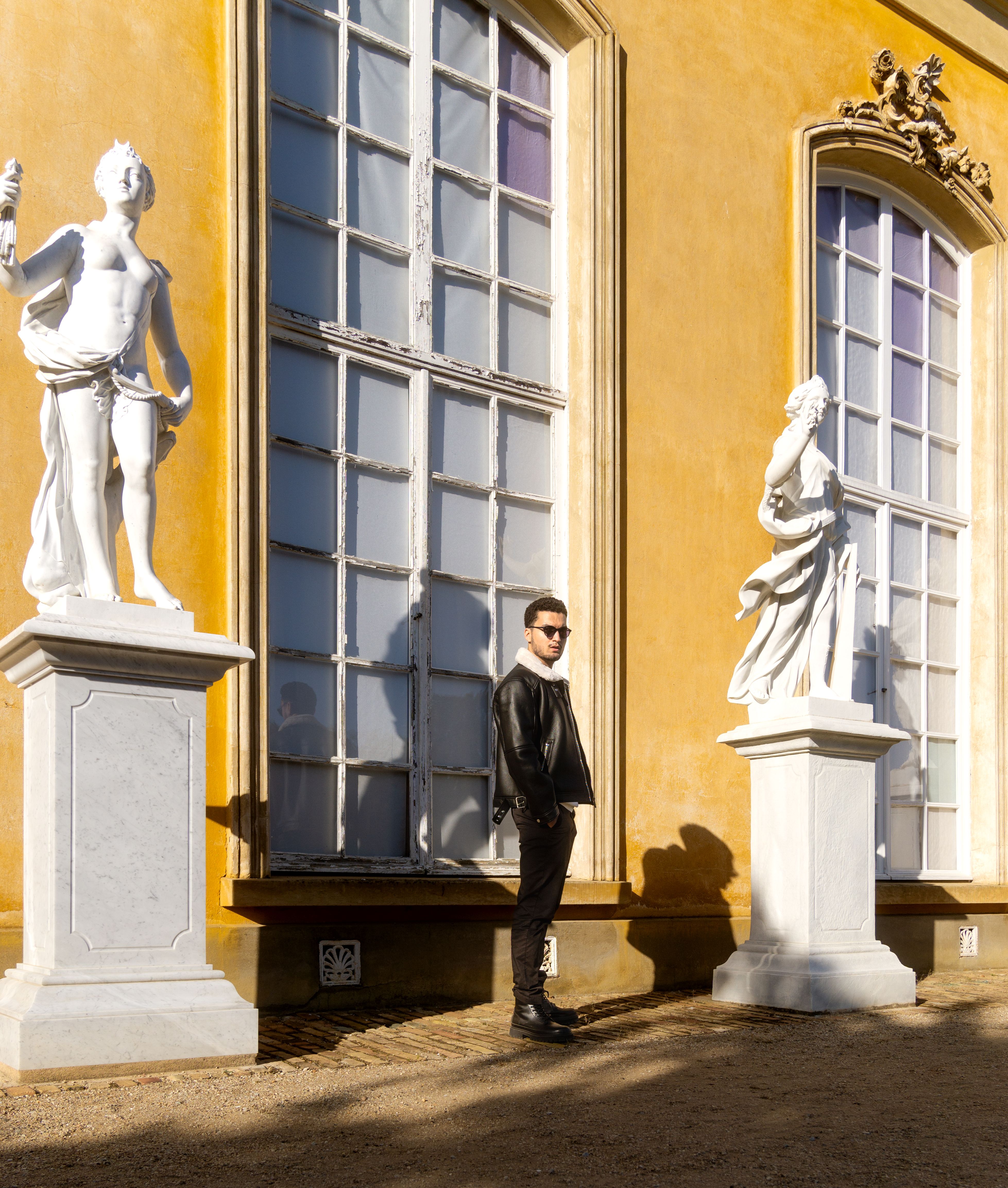 Man in sunglasses and leather jacket standing between classical statues in Potsdam, Germany.