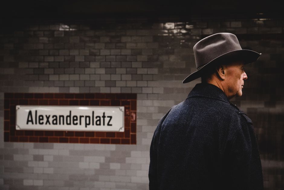 A man with a cowboy hat stands beside the Alexanderplatz subway sign in Berlin.