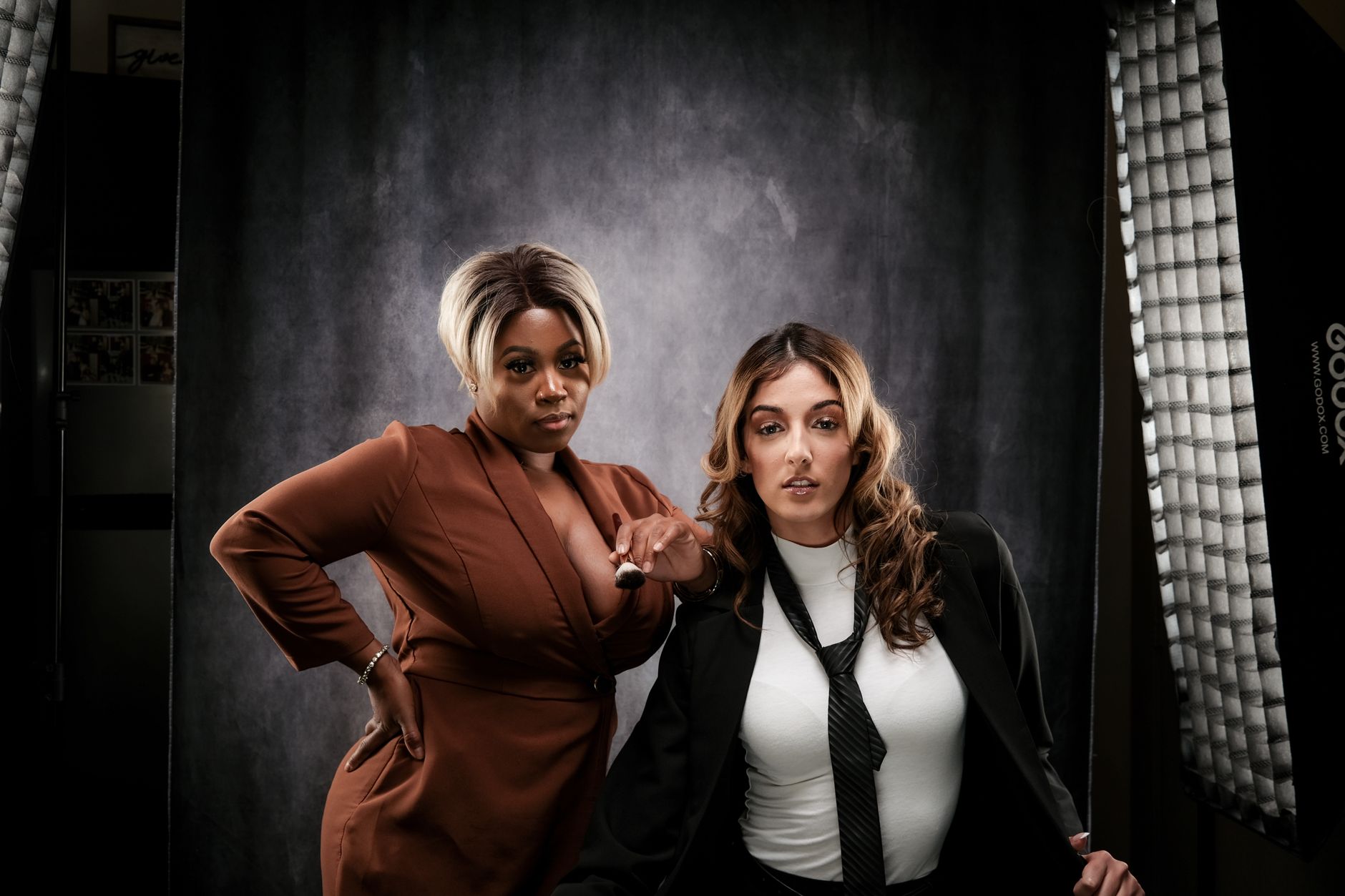 Two women pose confidently in professional attire in a studio setting.