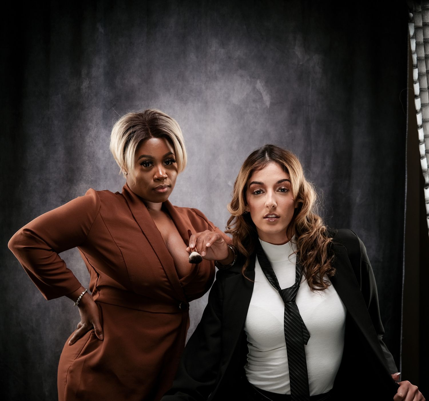 Two women pose confidently in professional attire in a studio setting.