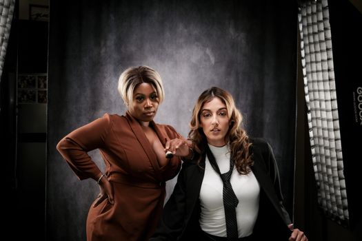 Two women pose confidently in professional attire in a studio setting.