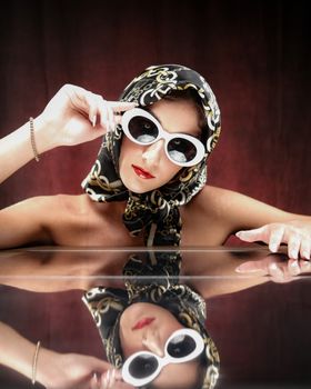 Fashionable woman with headscarf and sunglasses mirrored on a reflective surface.