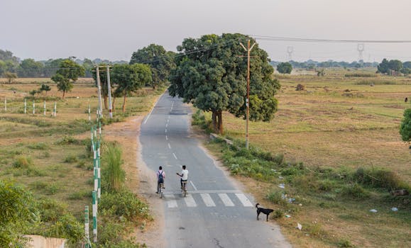 Two boys cycling on a rural road in Deeg, Uttar Pradesh, India with a black dog nearby.