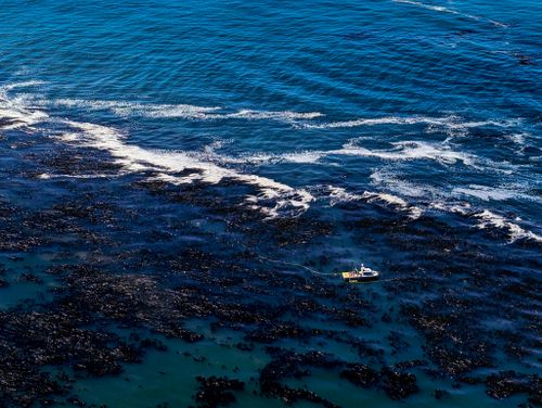 Free A small boat navigates through deep blue waters near a rugged coastline, viewed from above. Stock Photo
