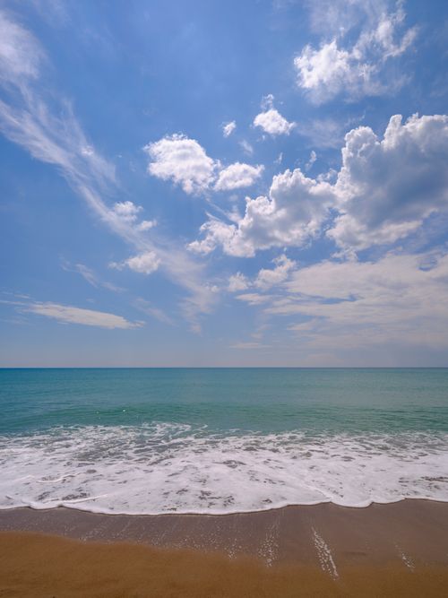 Free Peaceful beach scene with gentle waves, clear blue sky, and airy clouds, perfect for relaxation and travel inspiration. Stock Photo