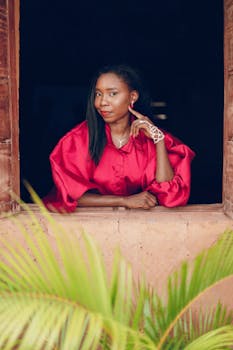 Elegant portrait of a woman in a vibrant red outfit framed by a rustic window, Nigeria.