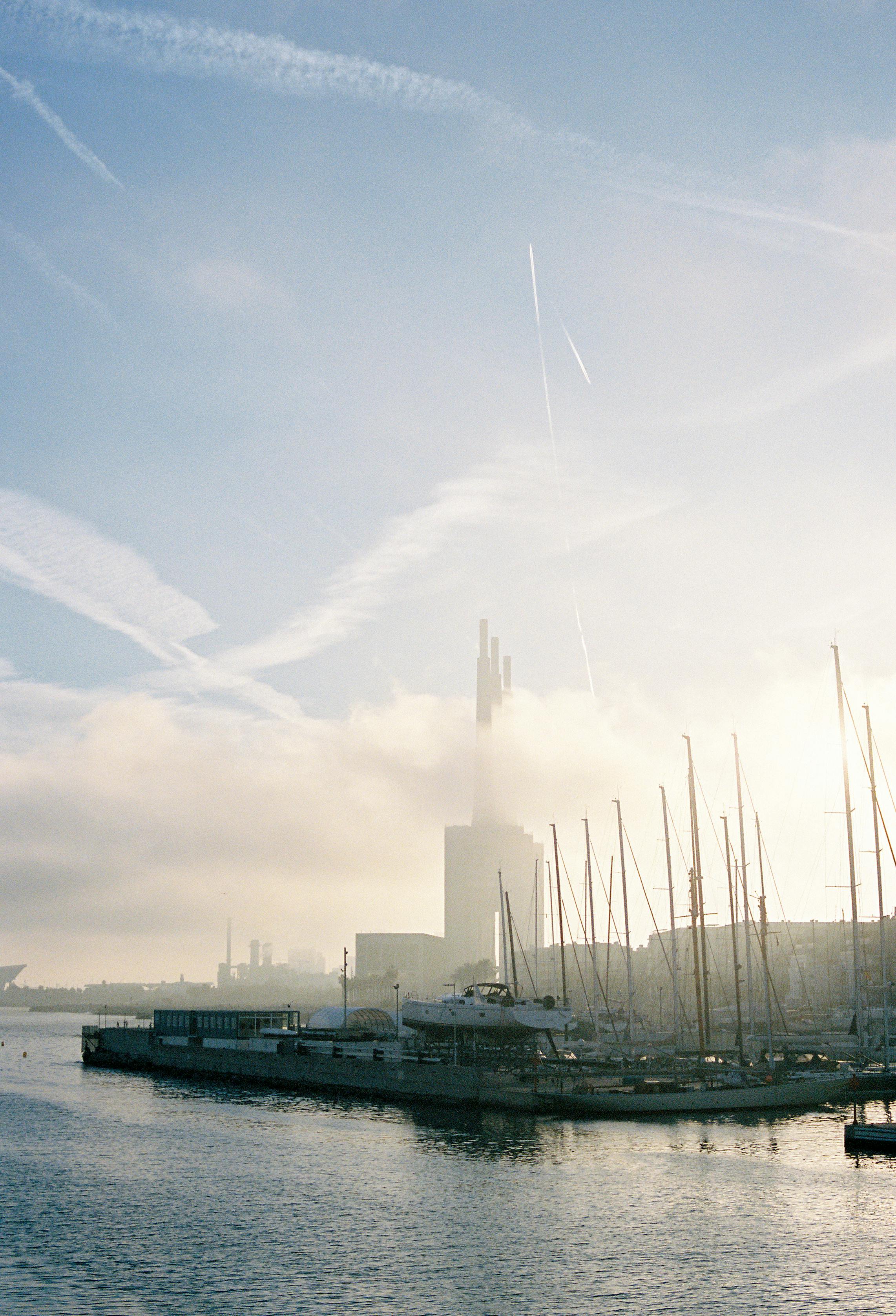 Marina at sunrise with tall ship masts and city skyline enveloped in mist, evoking tranquility.