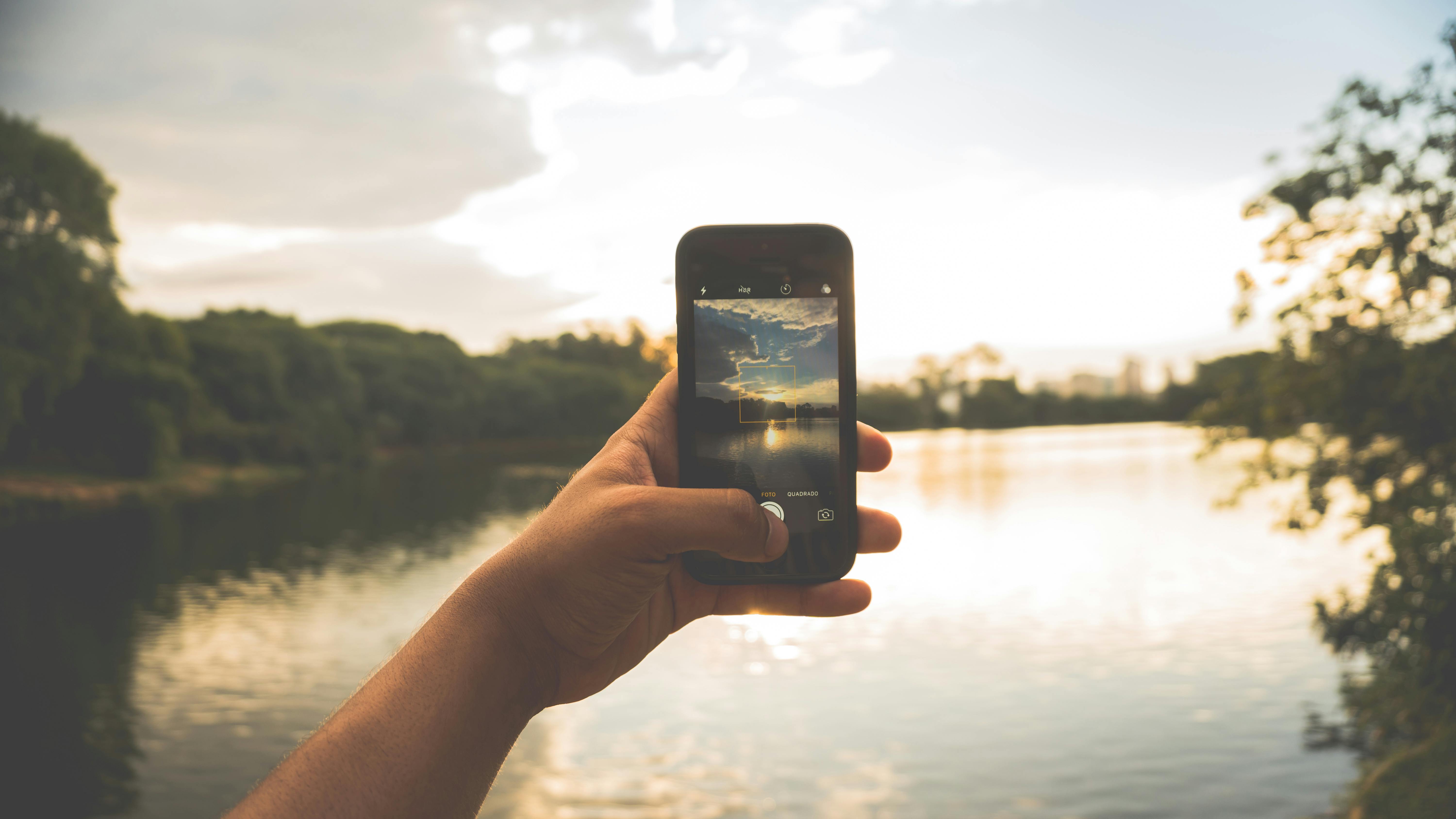 Hand photographing peaceful lake scenery with a smartphone at sunset