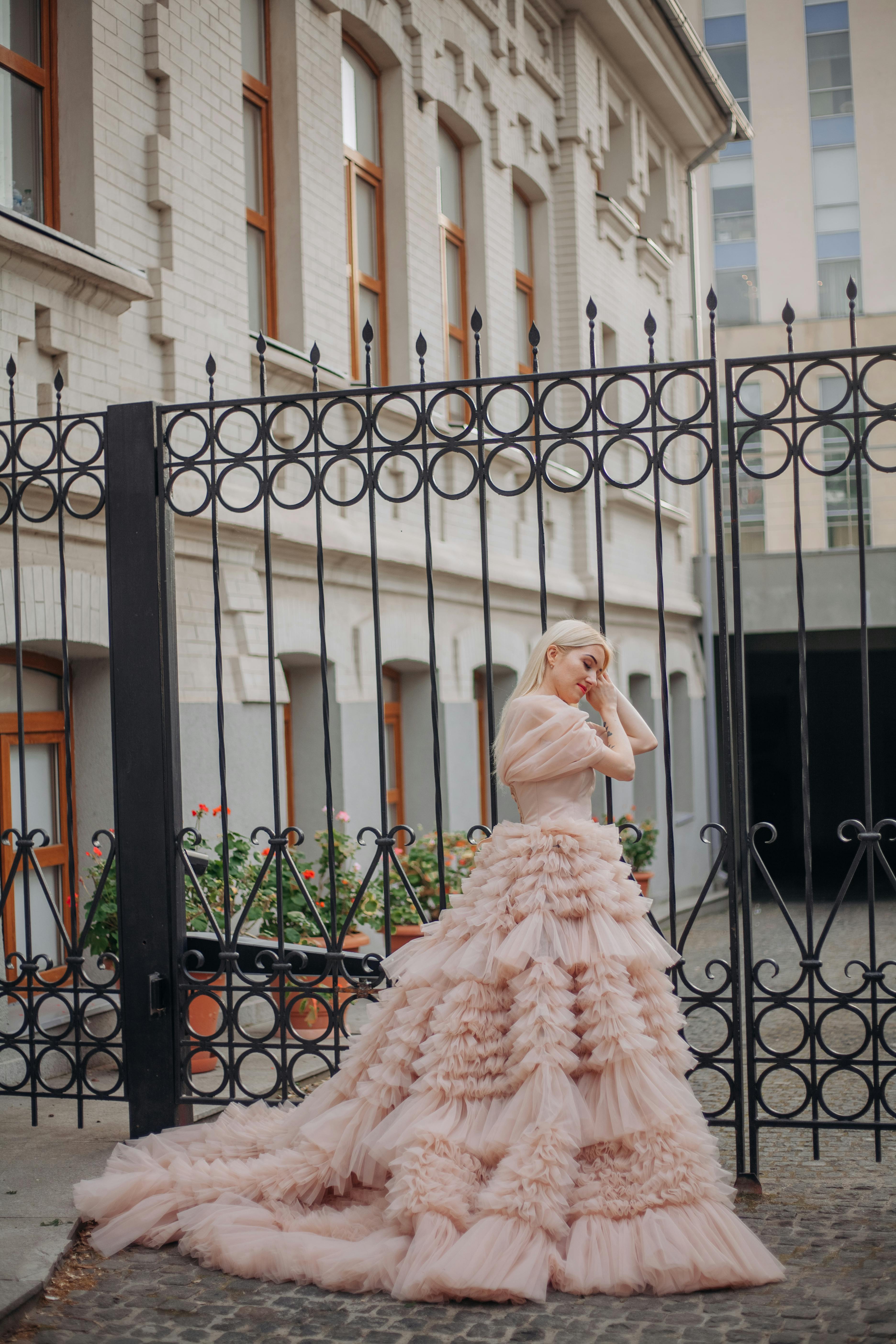 Free A woman in a luxurious pink gown poses gracefully by an ornate iron fence. Stock Photo