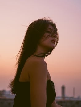 A young woman stands at sunset, hair blowing gently in the wind, evoking a serene and artistic mood.