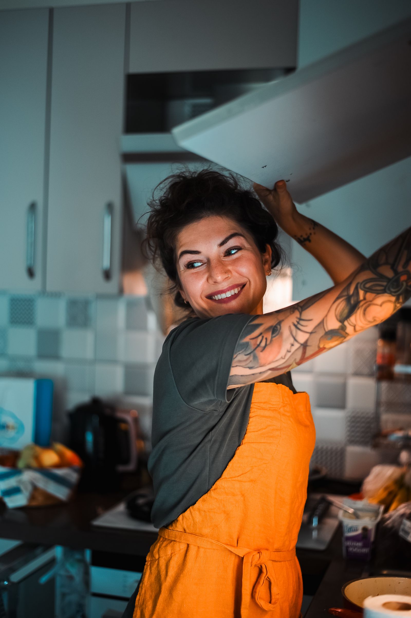 A joyful woman with tattoos preparing food in a cozy kitchen, wearing a bright orange apron.