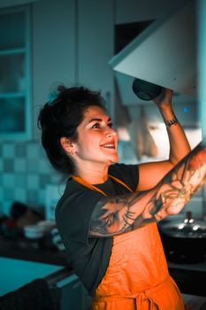 Tattooed woman in orange apron reaching into a kitchen cabinet with a smile, indoors.