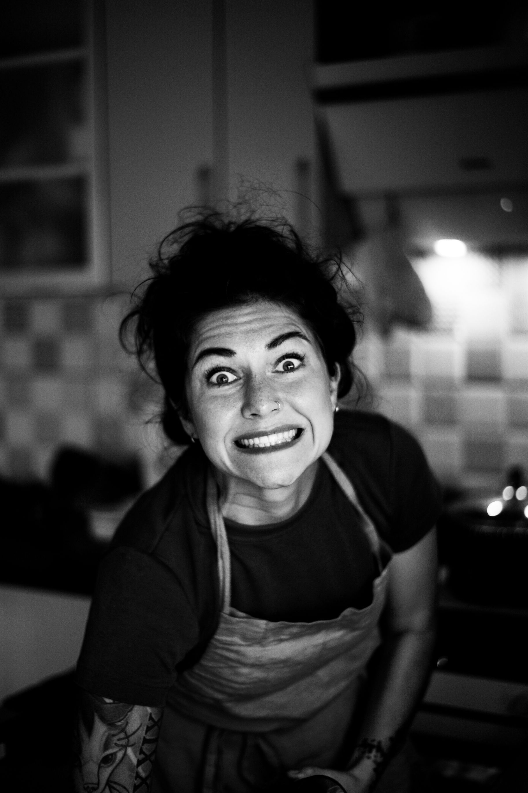 Black and white portrait of a woman showing a playful expression in a kitchen setting.