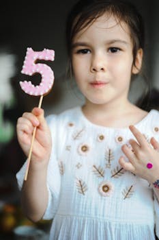 A young girl in a white dress holds a decorative number 5 stick to celebrate her birthday.