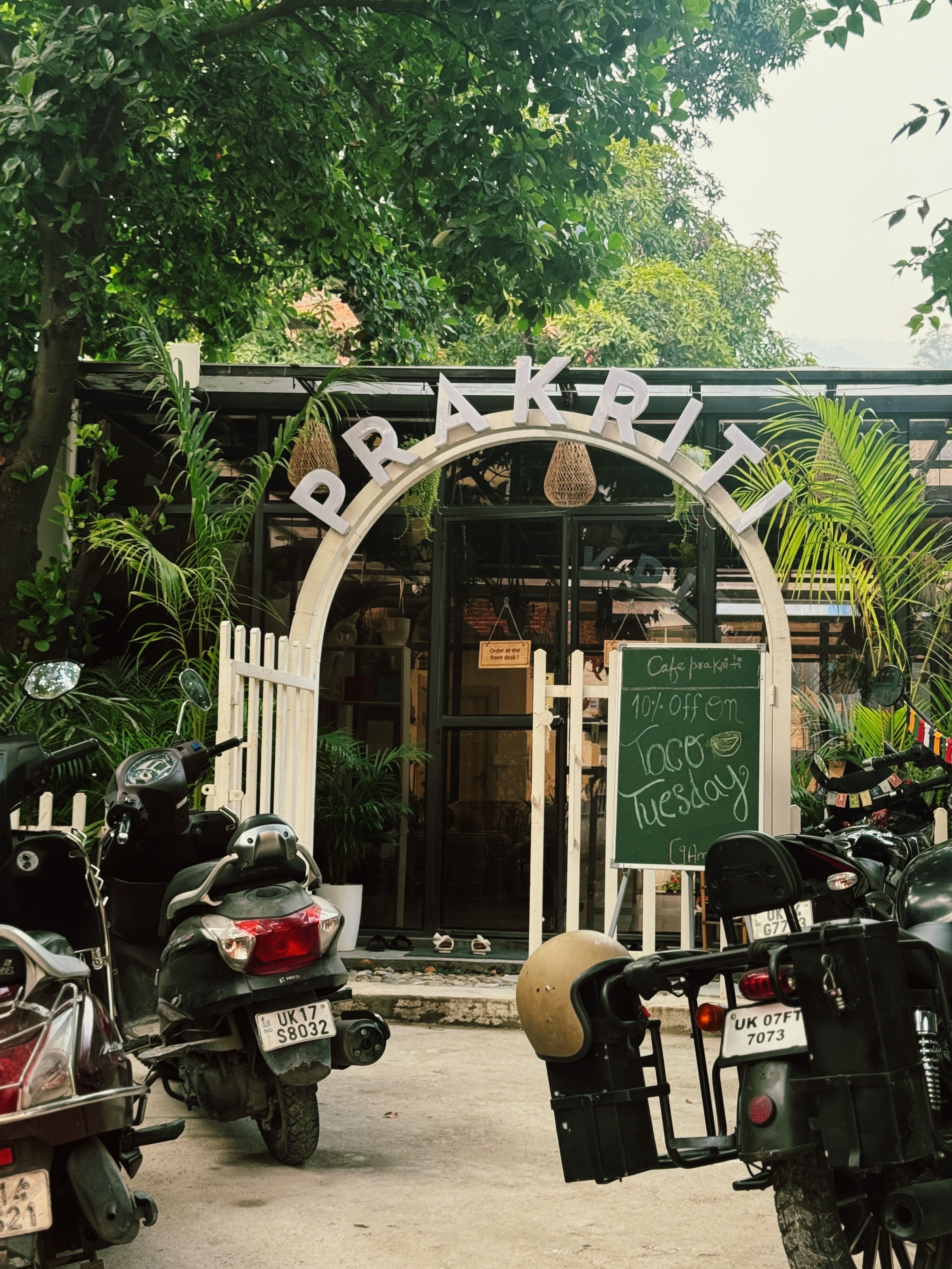 Cafe entrance with rustic sign, greenery, and parked scooters.