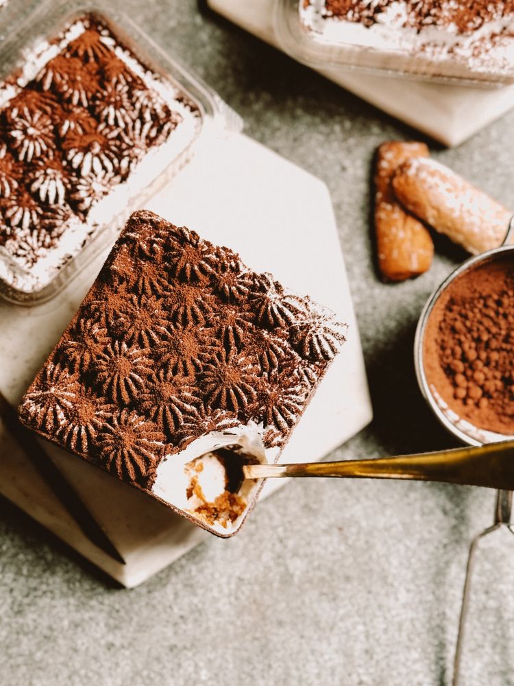Individual square tiramisu cups layered with espresso-soaked ladyfingers and white mascarpone cream beneath piped cocoa-dusted rosettes