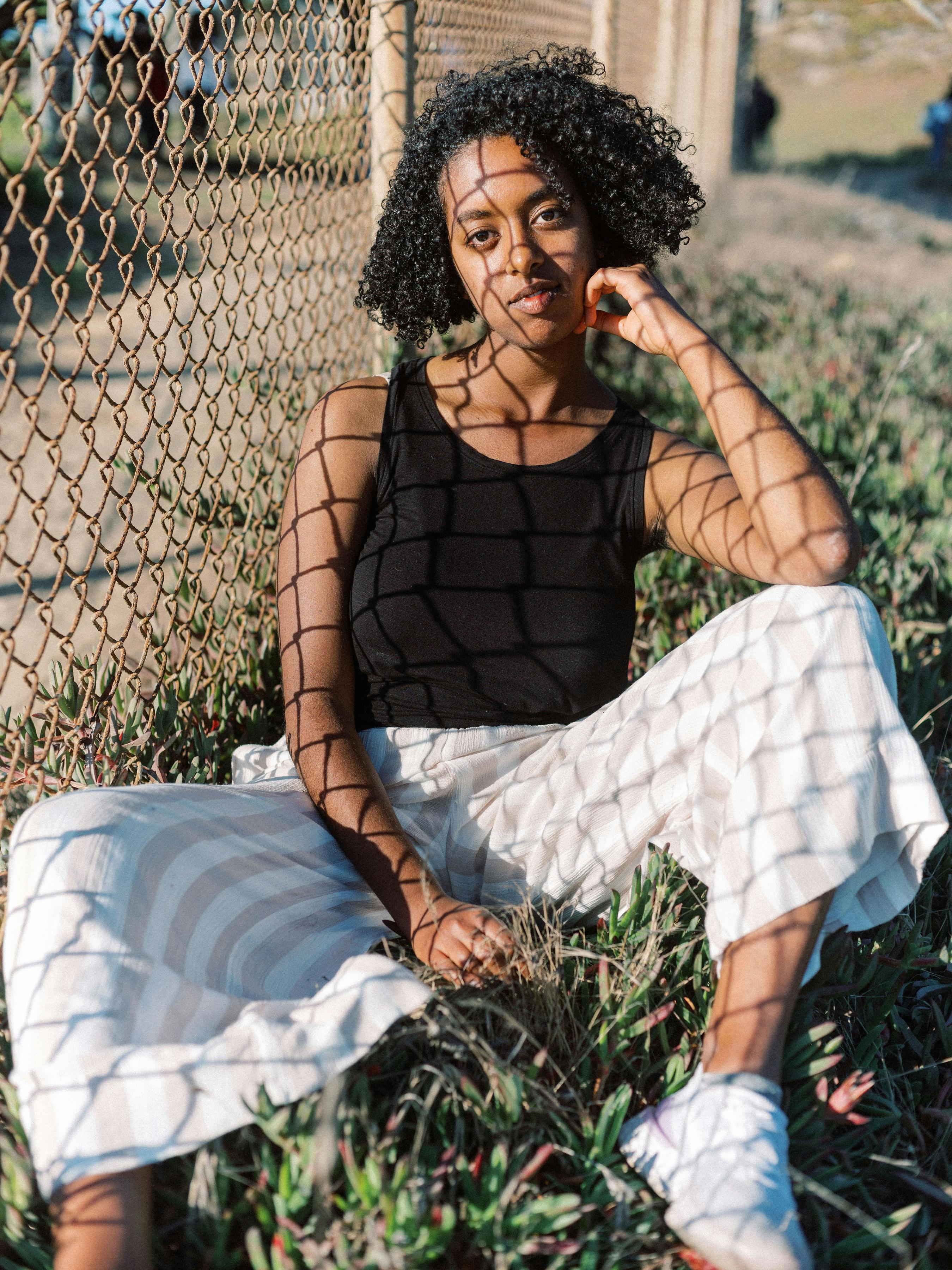 一位卷发的时尚女性坐在阳光下的栅栏旁，身着休闲装，神态轻松