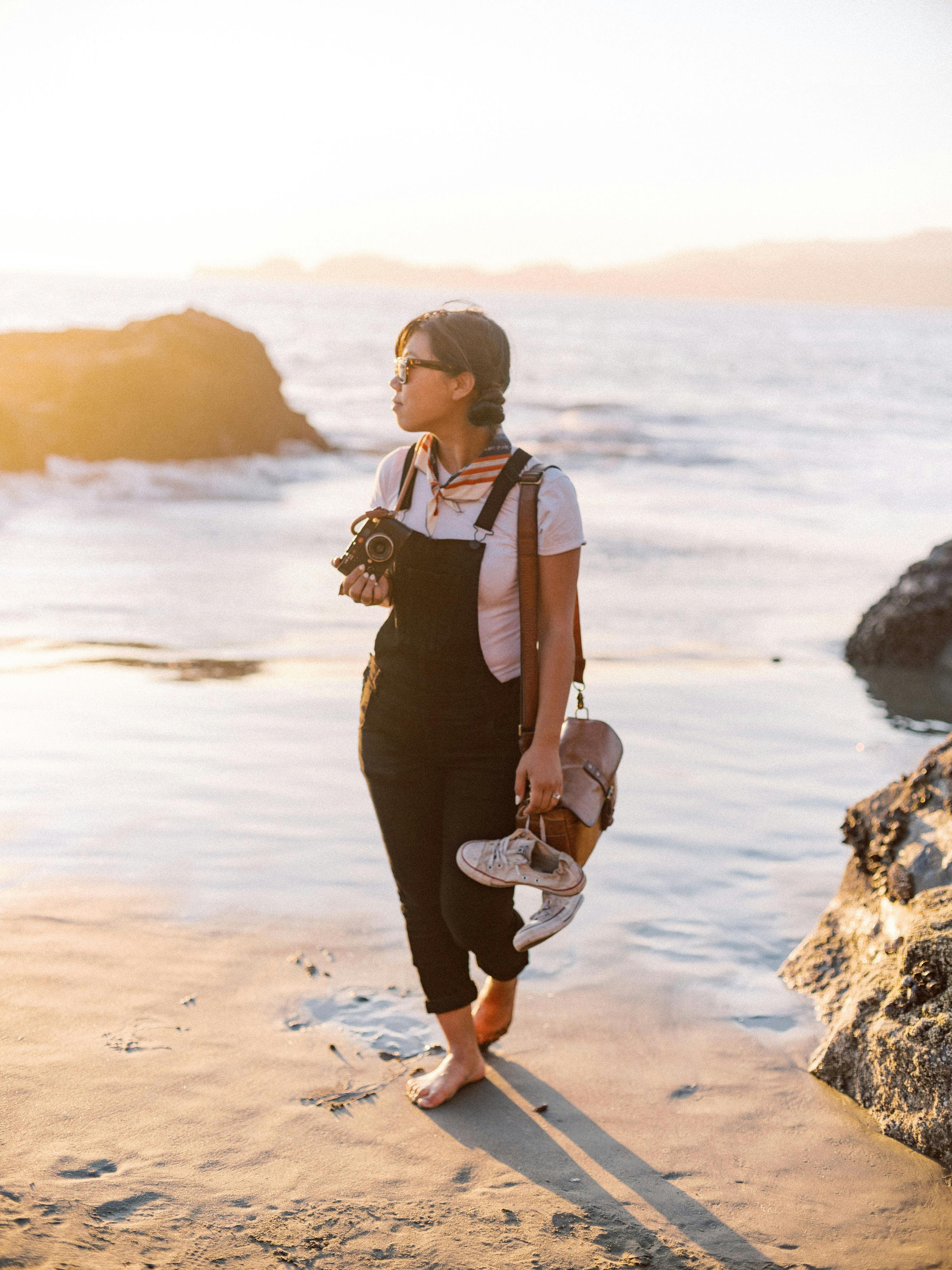 日落时分，一位年轻女子拿着相机和鞋子在海滩上漫步