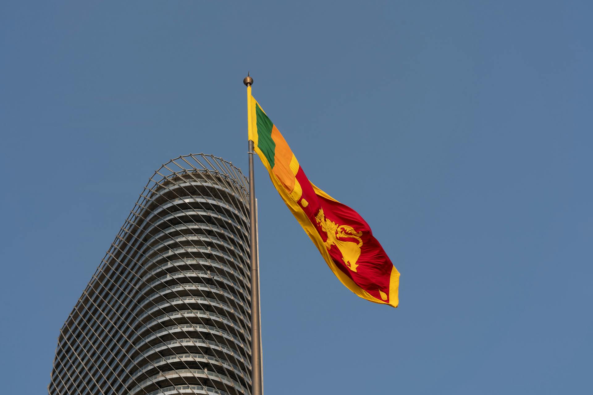 The Sri Lankan flag waving beside a modern Colombo skyscraper against a clear blue sky.