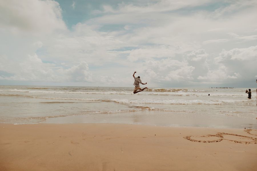 Person leaps joyfully on a sandy beach with waves and cloudy sky backdrop, capturing carefree summer vibes.