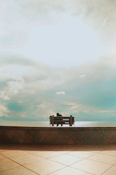 A solitary figure sits on a bench gazing at the vast ocean under a cloudy sky.