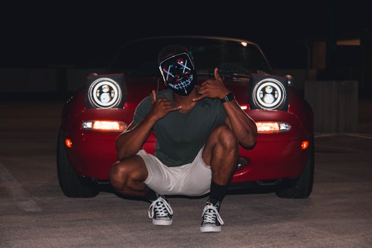 Man Wearing Mask Crouching In Front Of Red Car