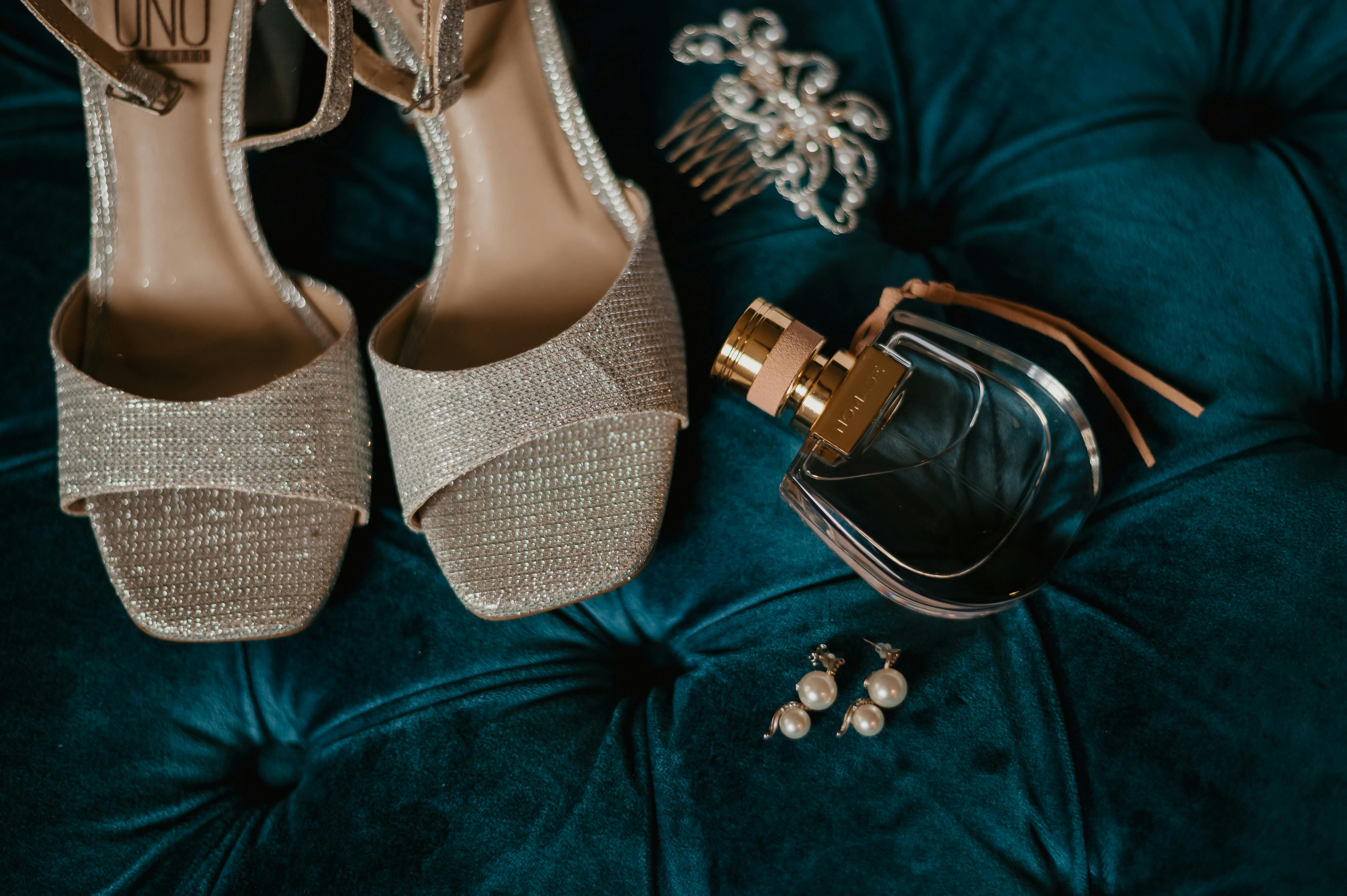 Chic arrangement of glittery heels, perfume, and jewelry on plush teal fabric.