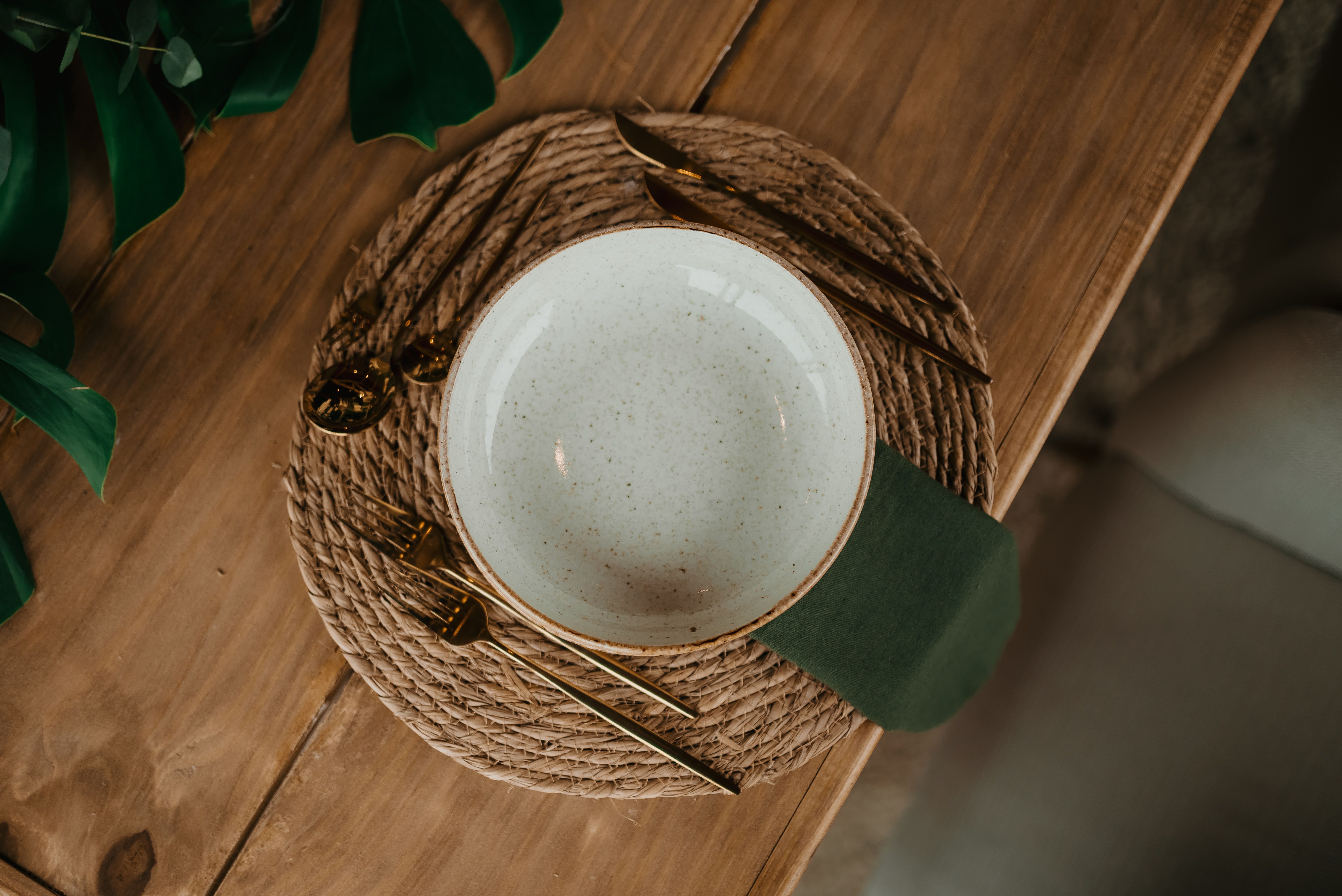 A speckled stoneware bowl with gold cutlery on a woven placemat and green linen napkin