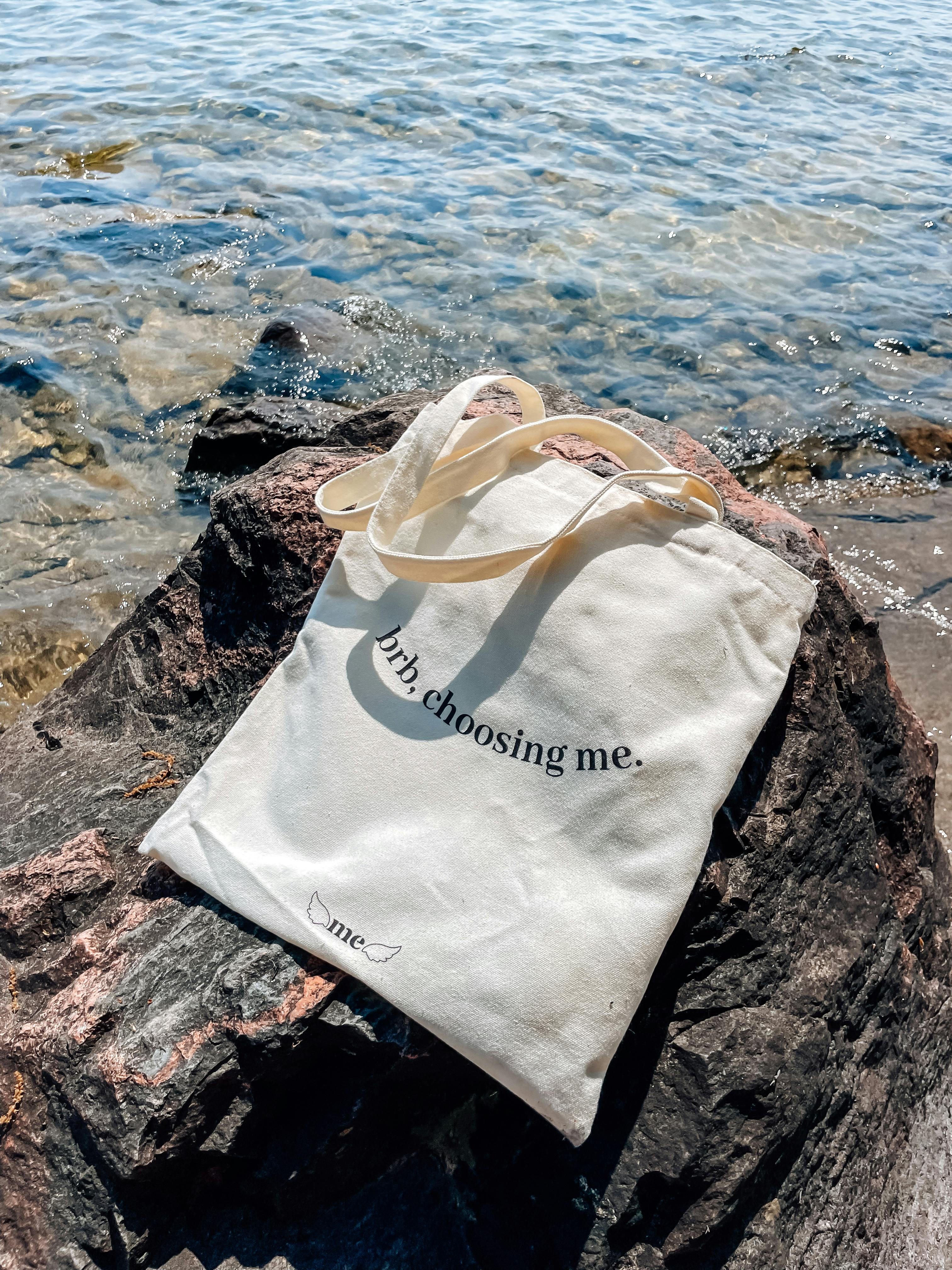 A canvas tote bag with text 'choosing me.' placed on rocky beach by the ocean.