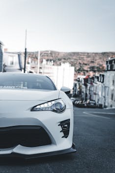 A modern white car parked on a steep urban street with houses and a distant hill in view.
