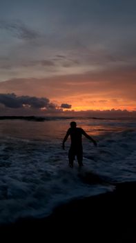 Silhouette of an individual standing in ocean waves against a vibrant sunset sky.