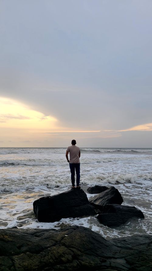 Free Man stands on rocks by the ocean, gazing at waves under a serene sunset sky. Stock Photo