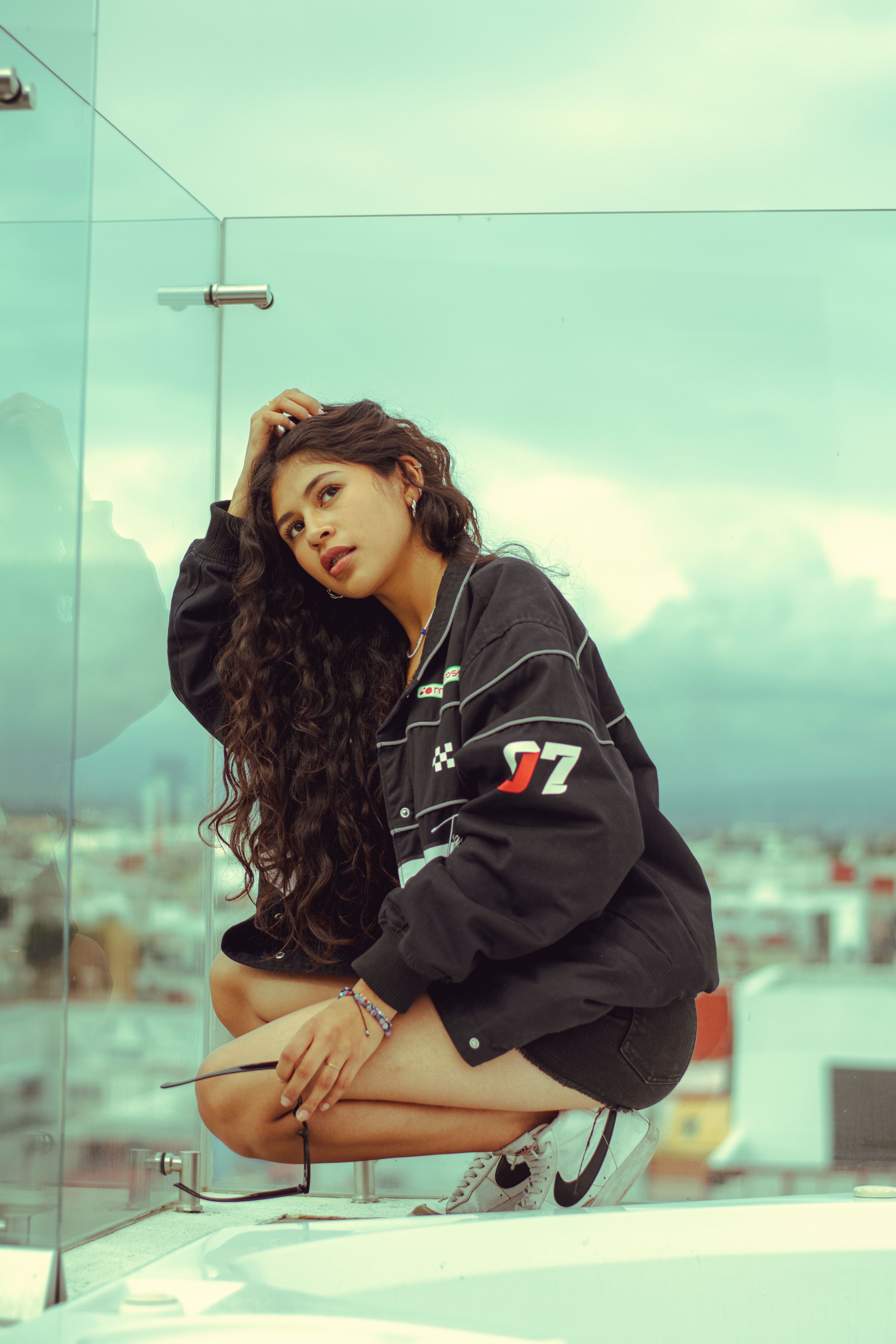 A woman squats on a rooftop with urban skyline in the background, featuring cloudy sky.