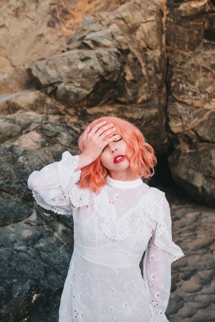 Stylish woman in white dress poses with hand on face against rocky backdrop.