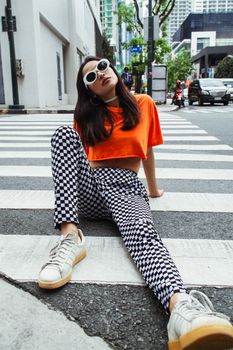 Fashionable woman in orange top and checkered pants posing on a city street crosswalk.