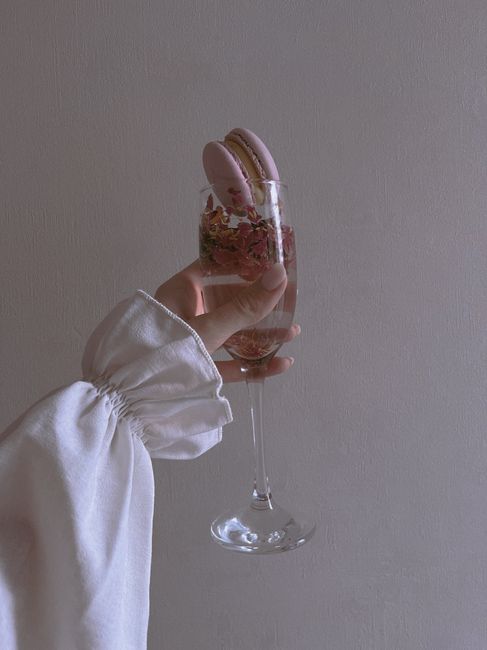 A hand holding a champagne glass with pink macarons and floral decorations, creating an artistic presentation.
