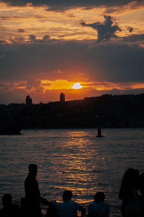 Free A dramatic sunset creating silhouettes of people by the water with a cityscape backdrop. Stock Photo