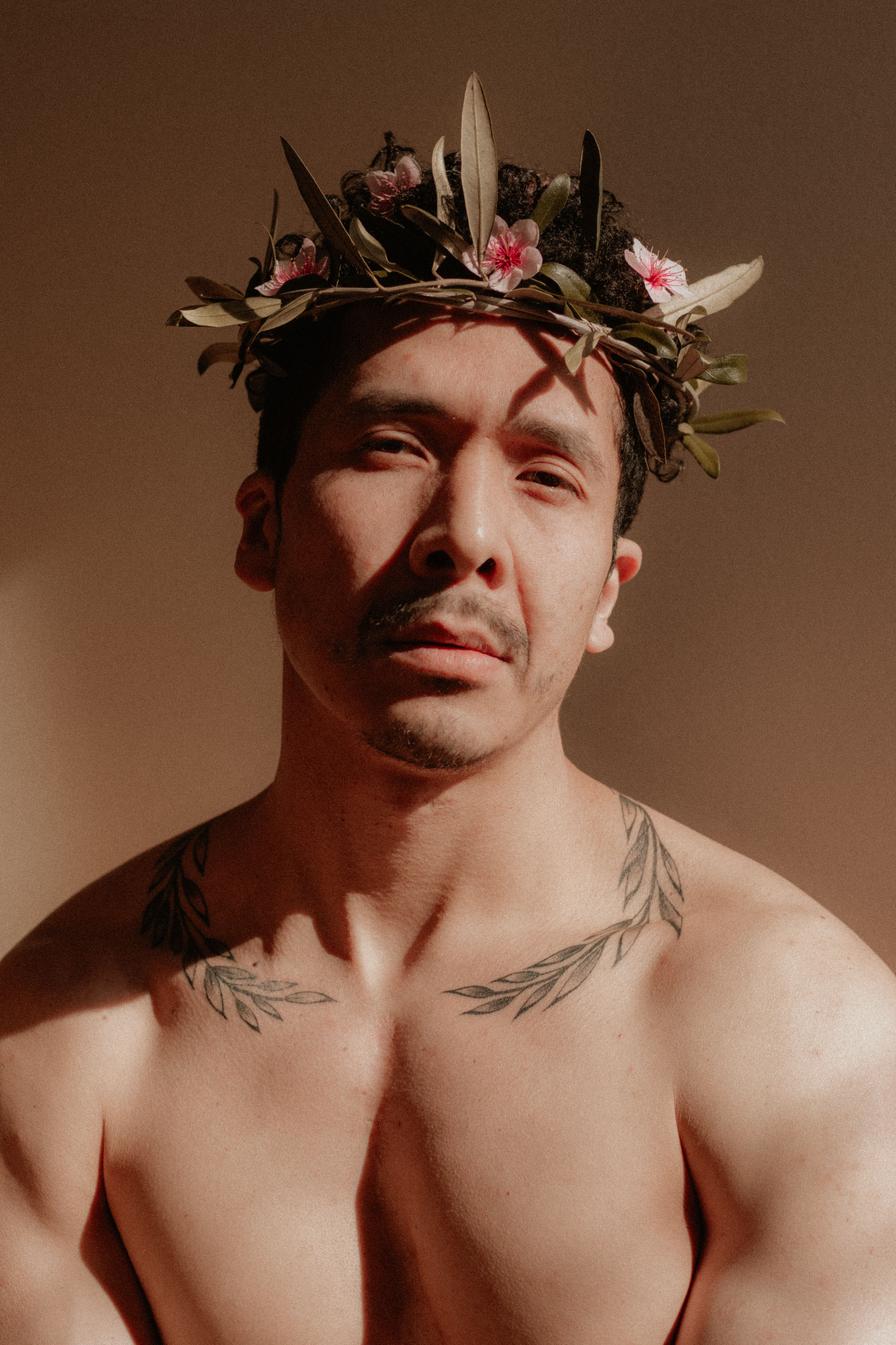 A tattooed man wearing a floral crown poses shirtless in warm sunlight, exuding strength and artistic charm.