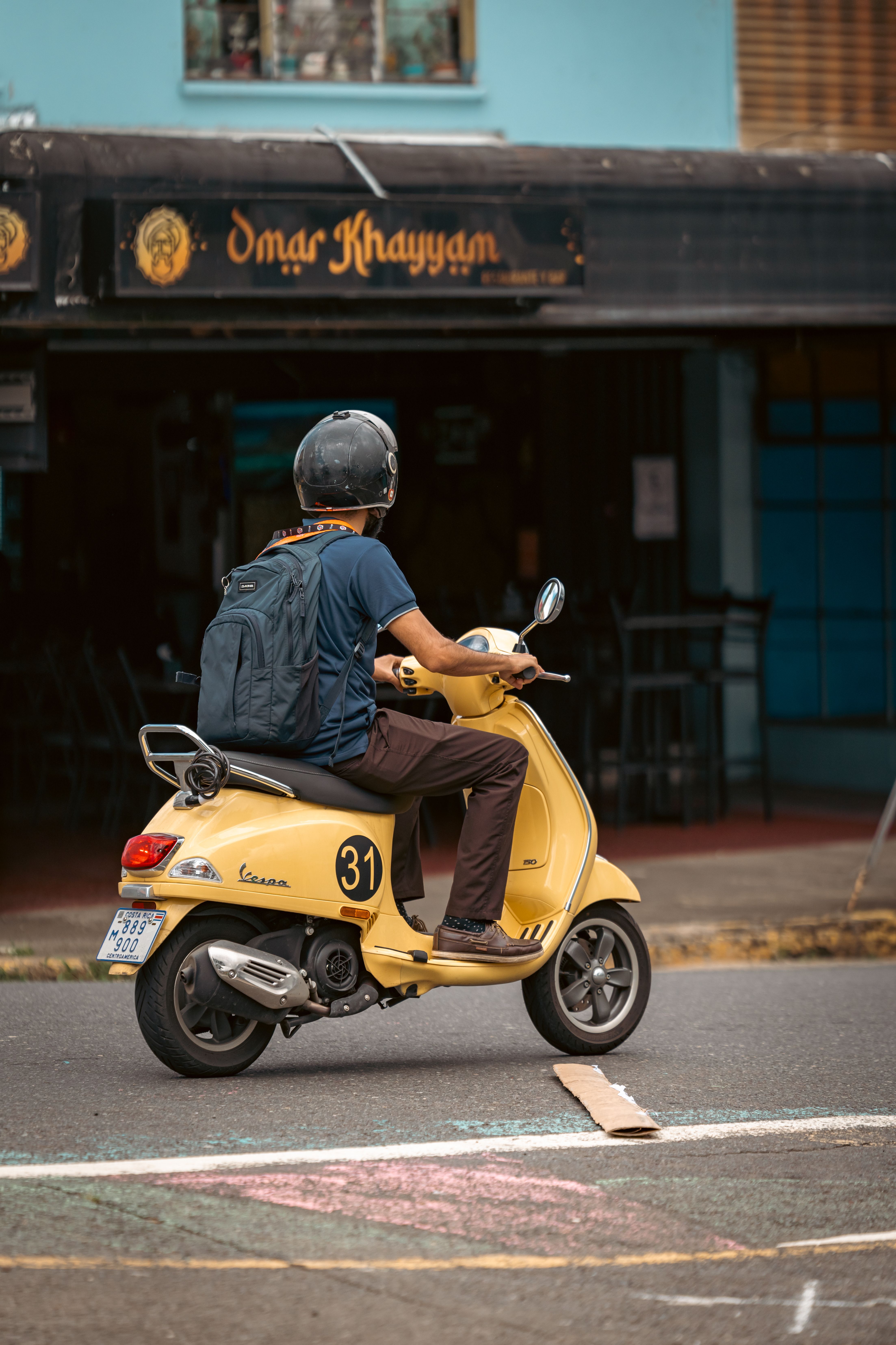 Personne conduisant un scooter jaune dans une rue urbaine, devant un café