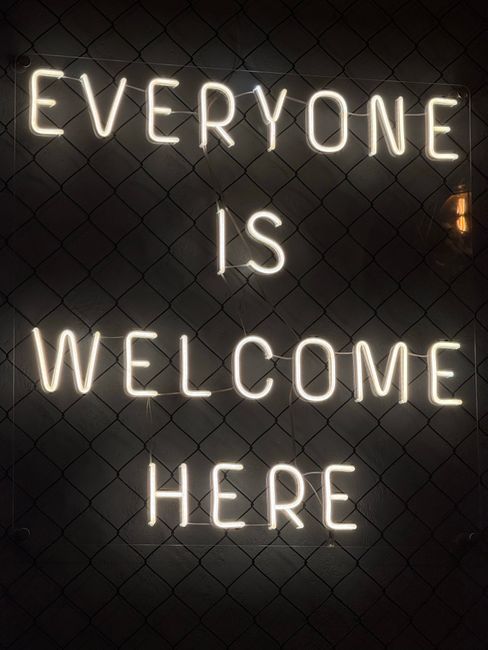 Illuminated neon sign reading 'Everyone is Welcome Here' on a wire fence.