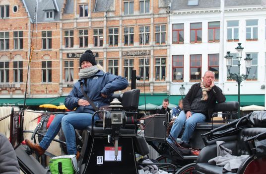 Horse carriage drivers resting in a historic square in Bruges, Belgium, showcasing local architecture and culture.