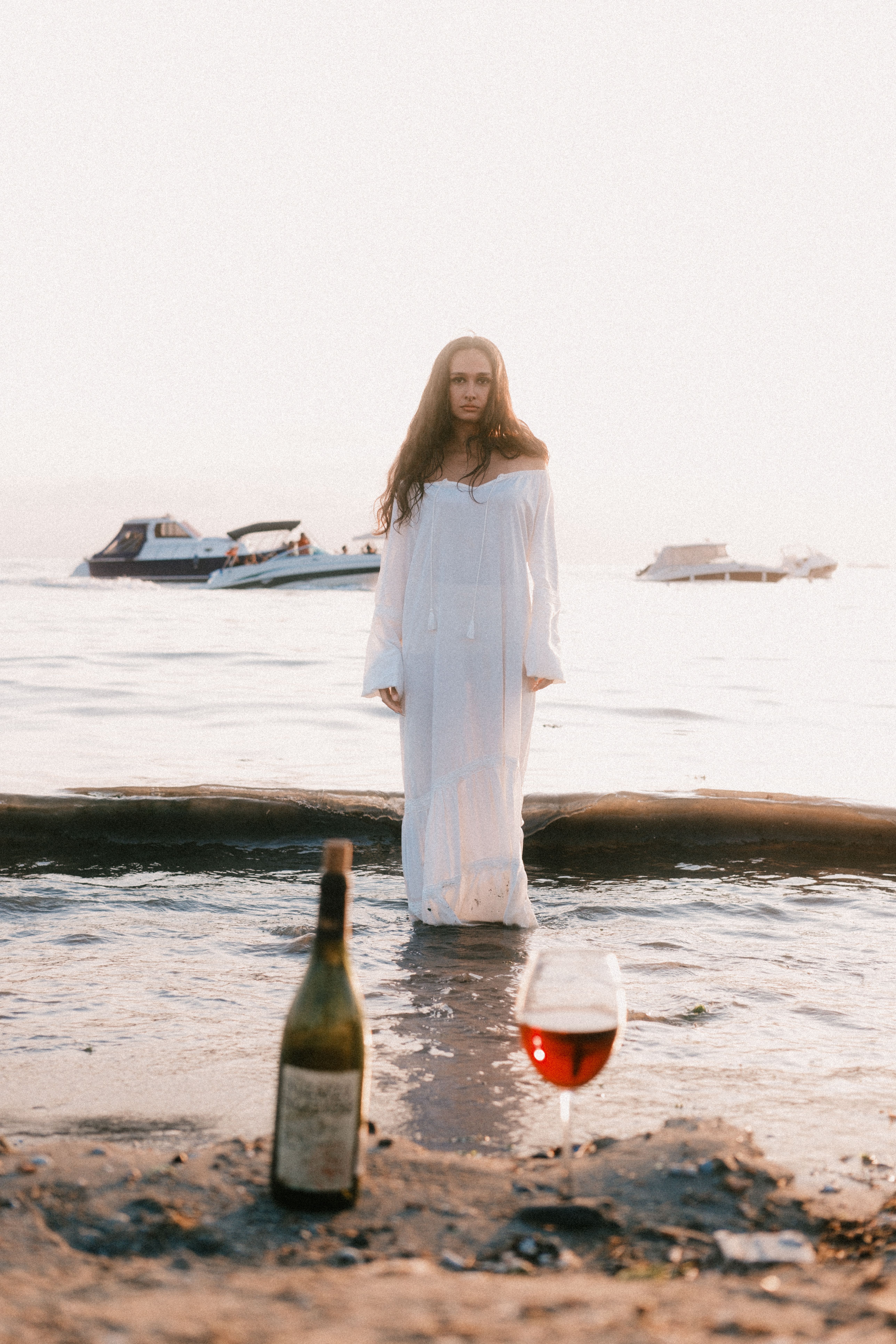 A mysterious woman in white stands on a beach with wine in foreground.