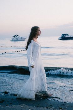 Serene image of a woman in a white dress walking by the sea at dusk with boats in the distance.
