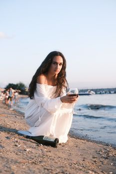 Woman in white dress enjoying a glass of wine on a beach during sunset.