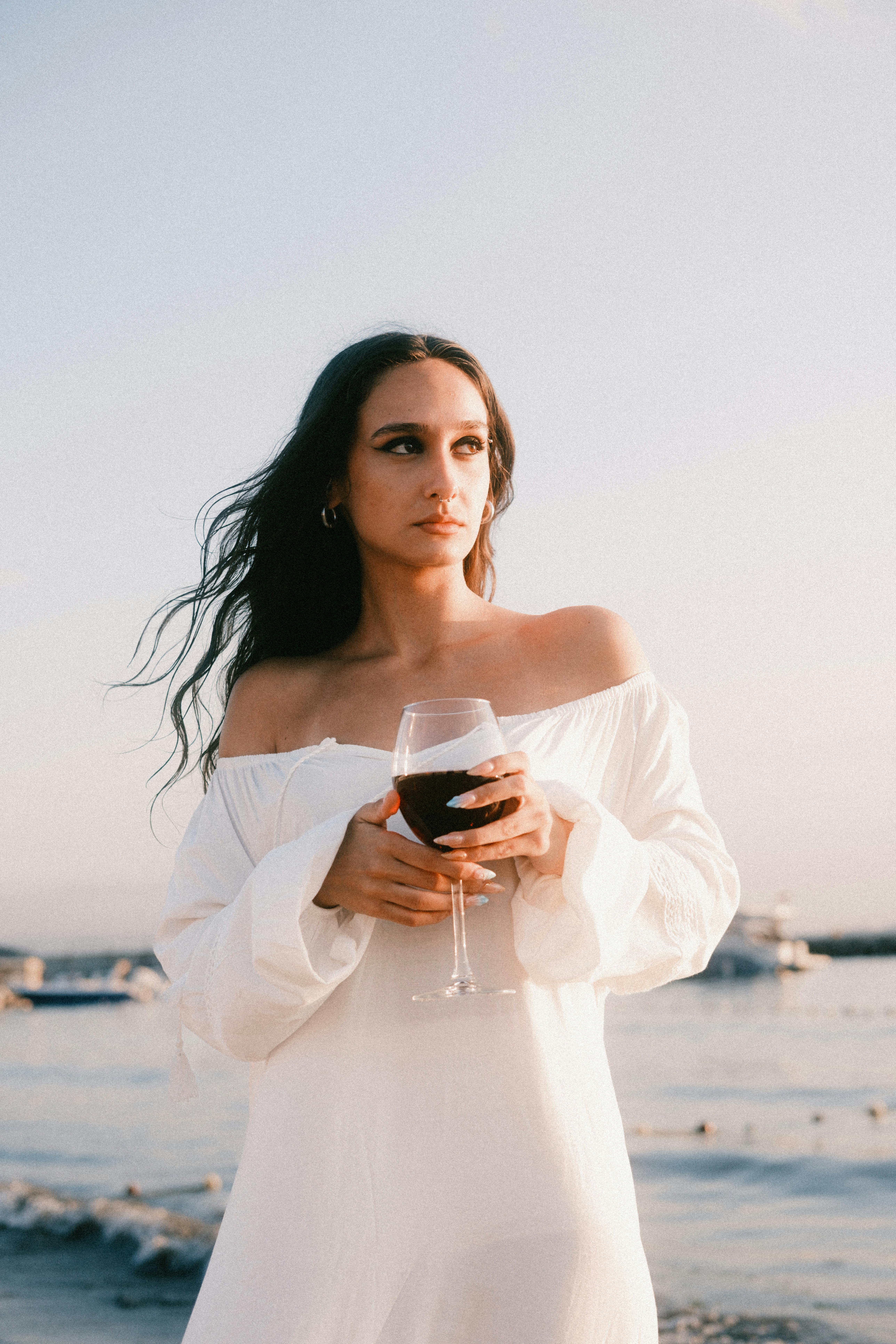 A tranquil scene of a woman in a white dress holding wine by the beach.