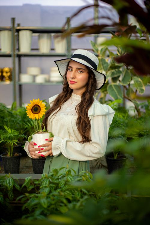 Free A young woman in a garden, wearing a hat, holding a potted sunflower amidst lush green plants. Stock Photo