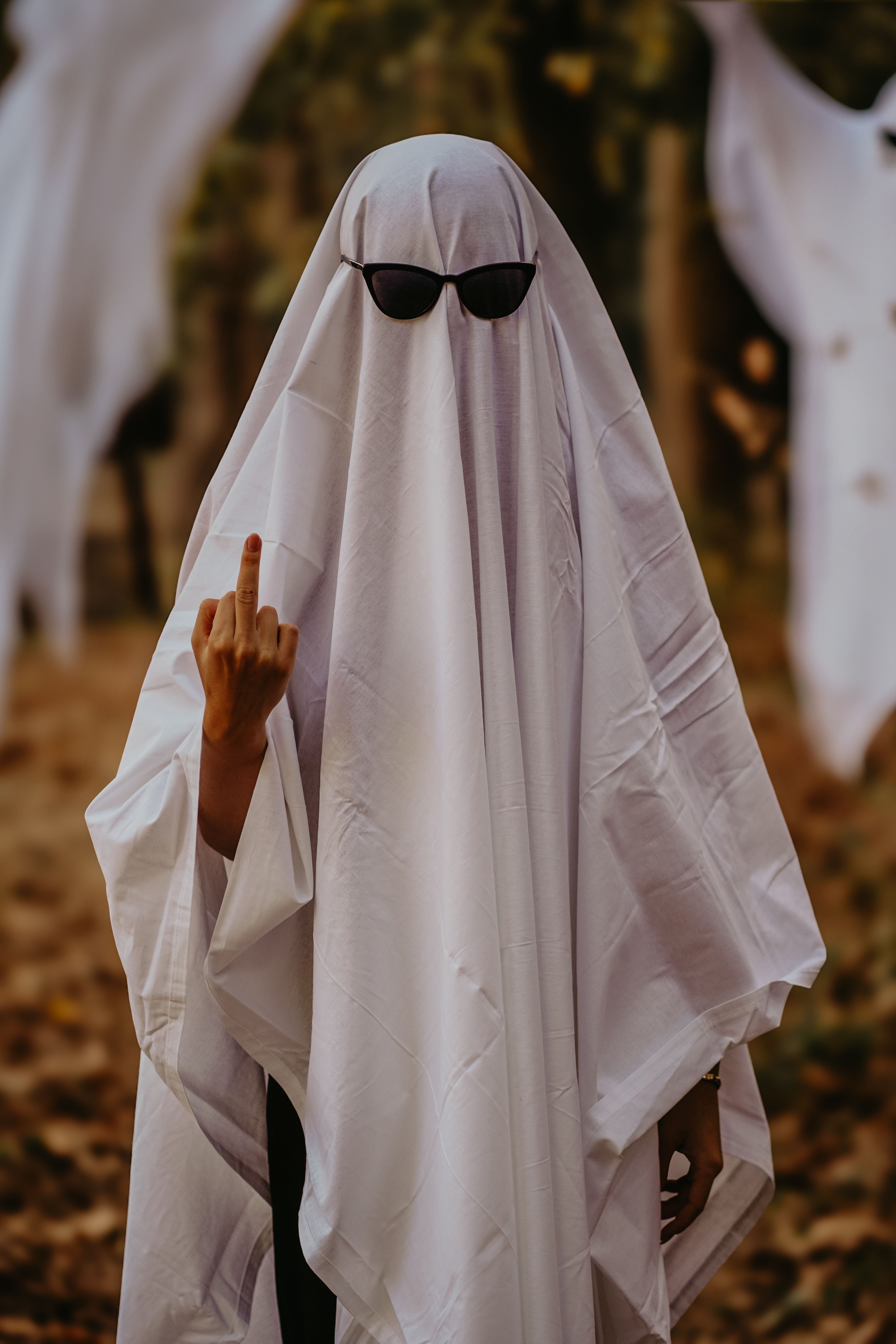Humorous ghost costume with sunglasses and hand gesture in autumn forest.
