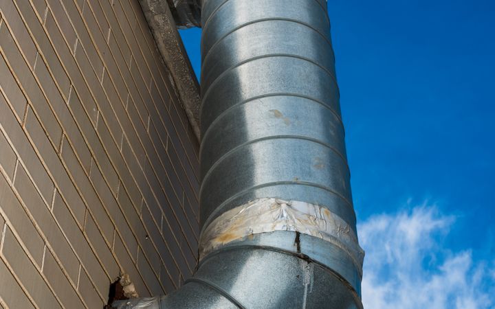 A large industrial air duct attached to a brick wall under a bright blue sky.